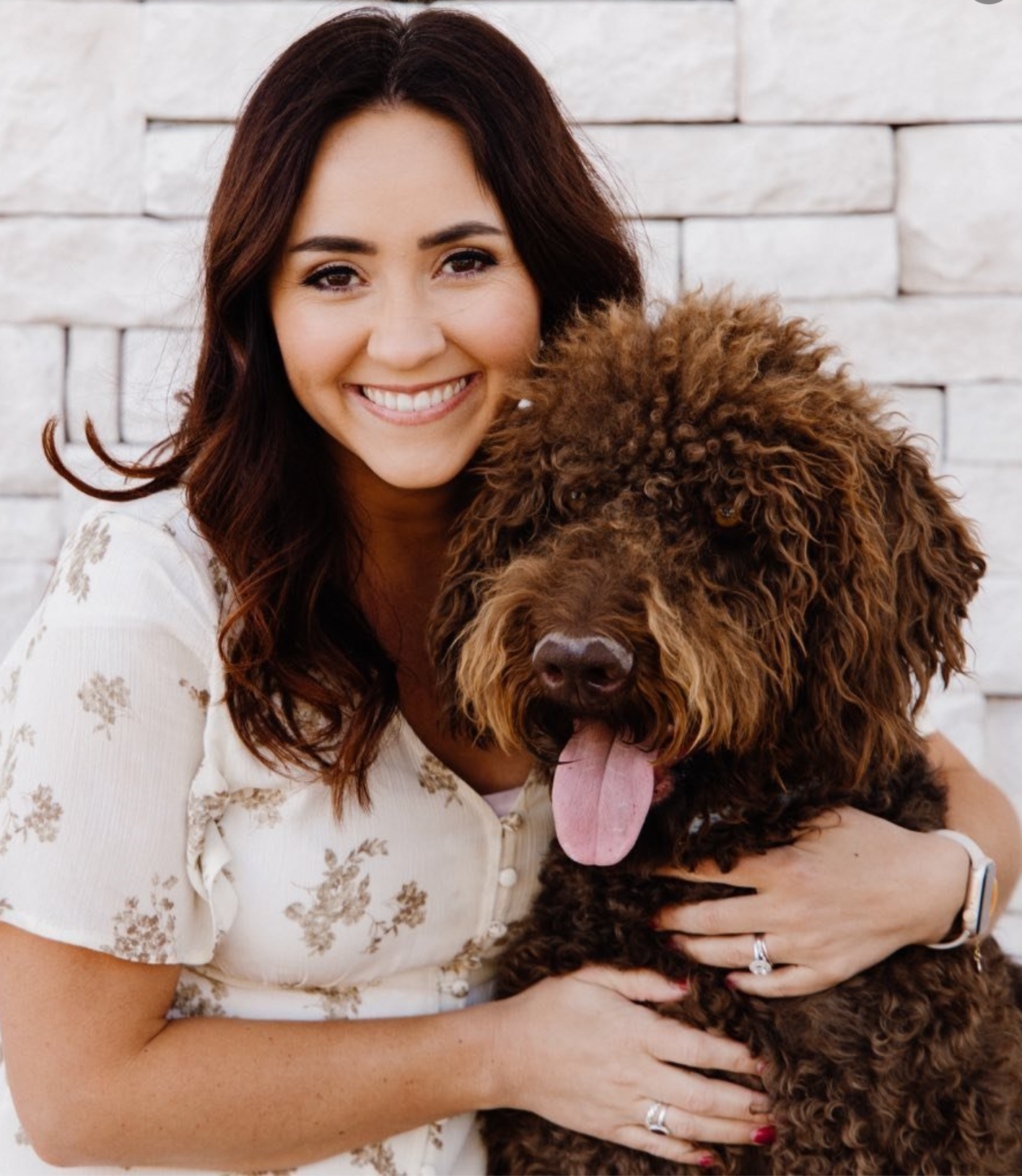 A fluffy brown doodle dog poses with someone against a white brick wall.