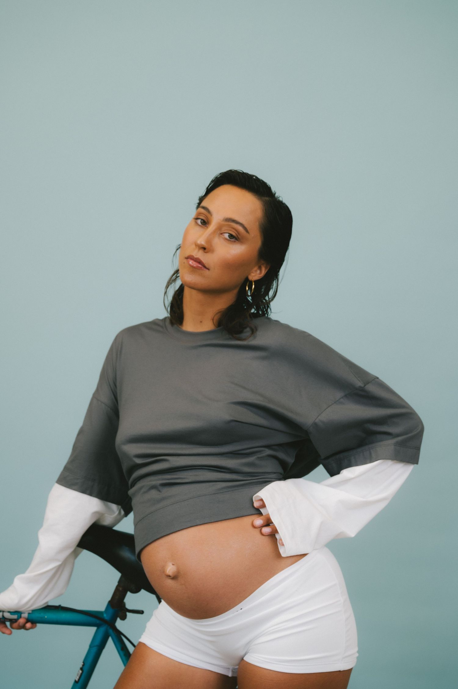 A pregnant woman in a grey sweatshirt and white shorts poses confidently next to a blue bicycle against a blue backdrop.