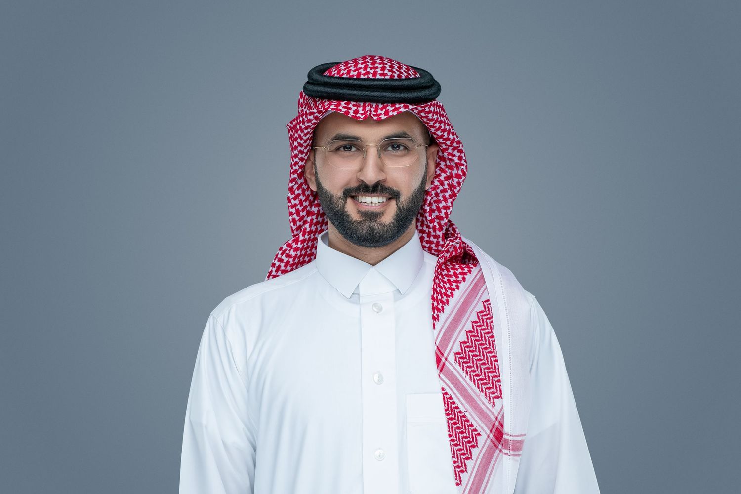 A person wearing traditional Saudi Arabian dress including white thobe and red and white keffiyeh against a gray background.