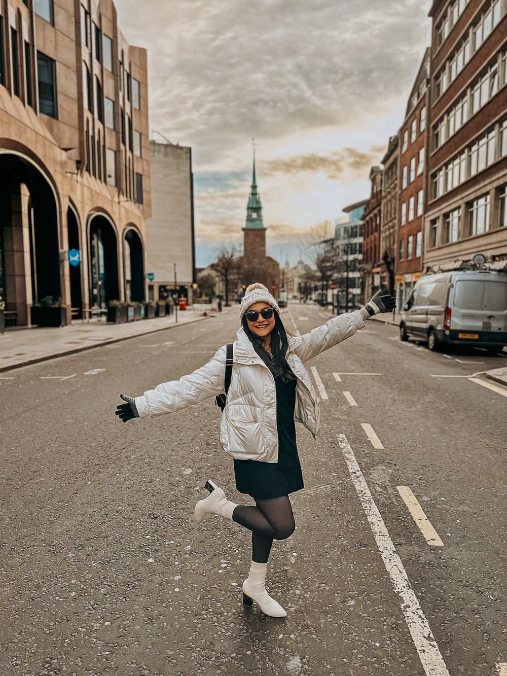 A person in winter attire strikes a joyful pose on a city street with historic architecture.