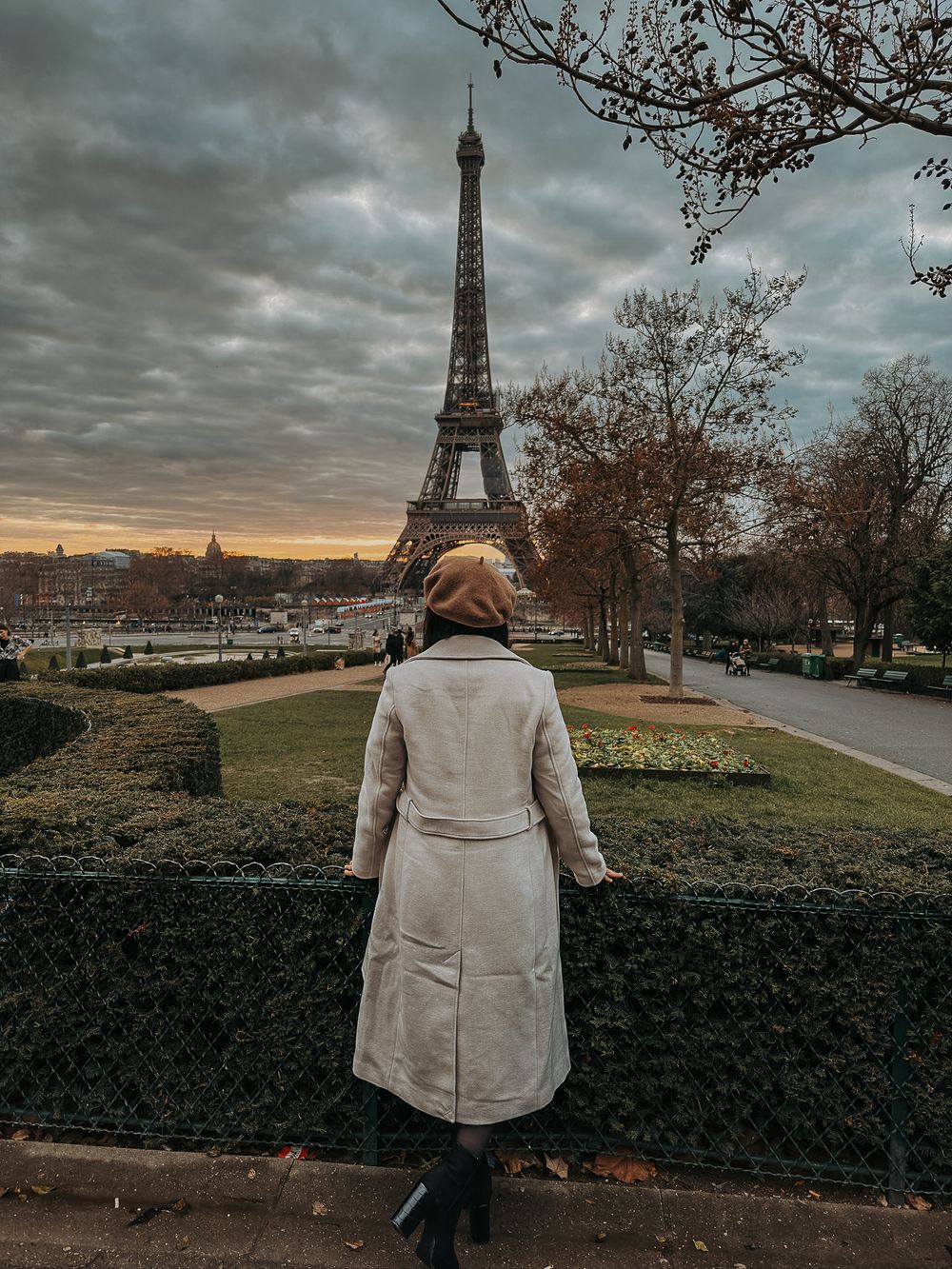 Person in beige coat admiring the Eiffel Tower at sunset against dramatic cloudy skies in Paris.