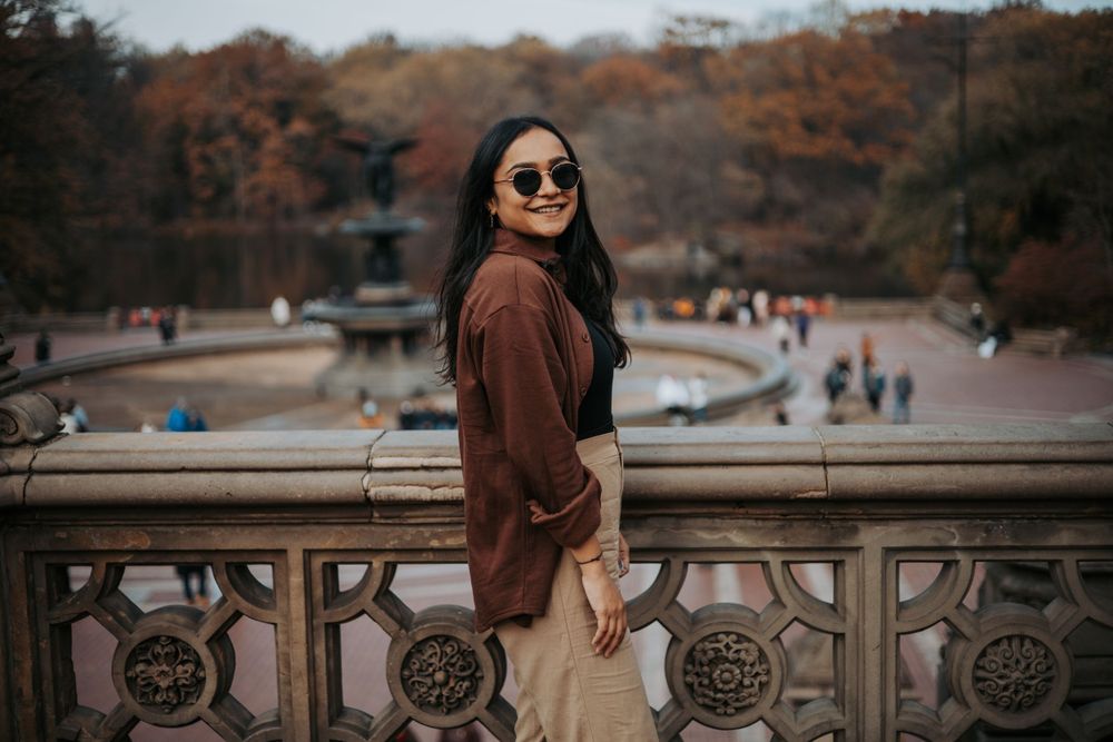 A person in sunglasses and brown jacket poses by an ornate railing with autumn scenery in background.