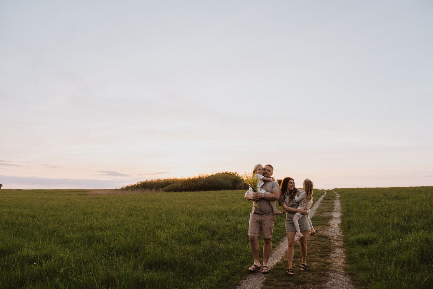 Two people stand together on a dirt path through a green field at sunset with soft pastel skies in the background.