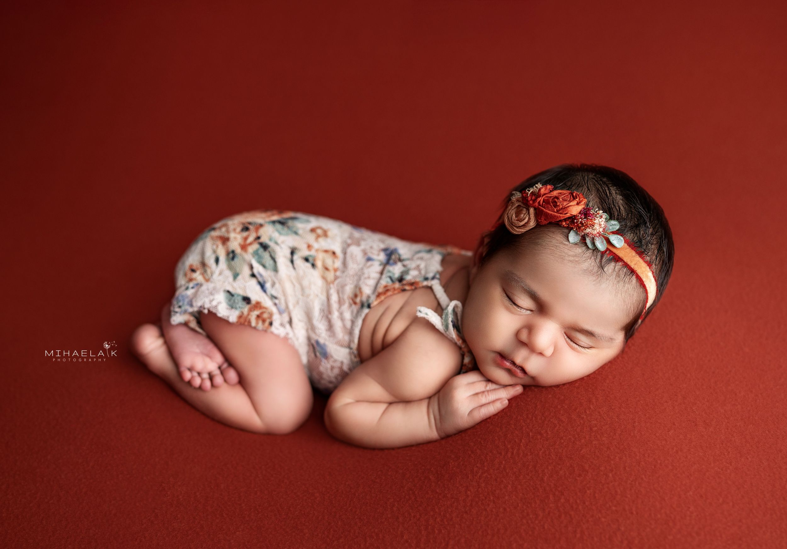 Newborn baby in rusty floral romper and headband on orange backdrop. Brooklyn NYC baby photography.