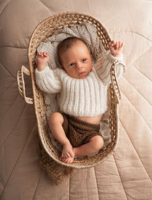 Newborn baby boy in neutral color knitted outfit posed in small rattan basket on neutral background, Brooklyn NYC newborn photographer.