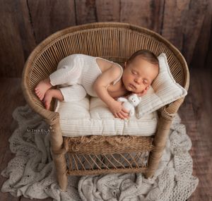 Newborn baby boy in white knitted outfit posed in rattan chair against woody background, Brooklyn NYC newborn photographer.