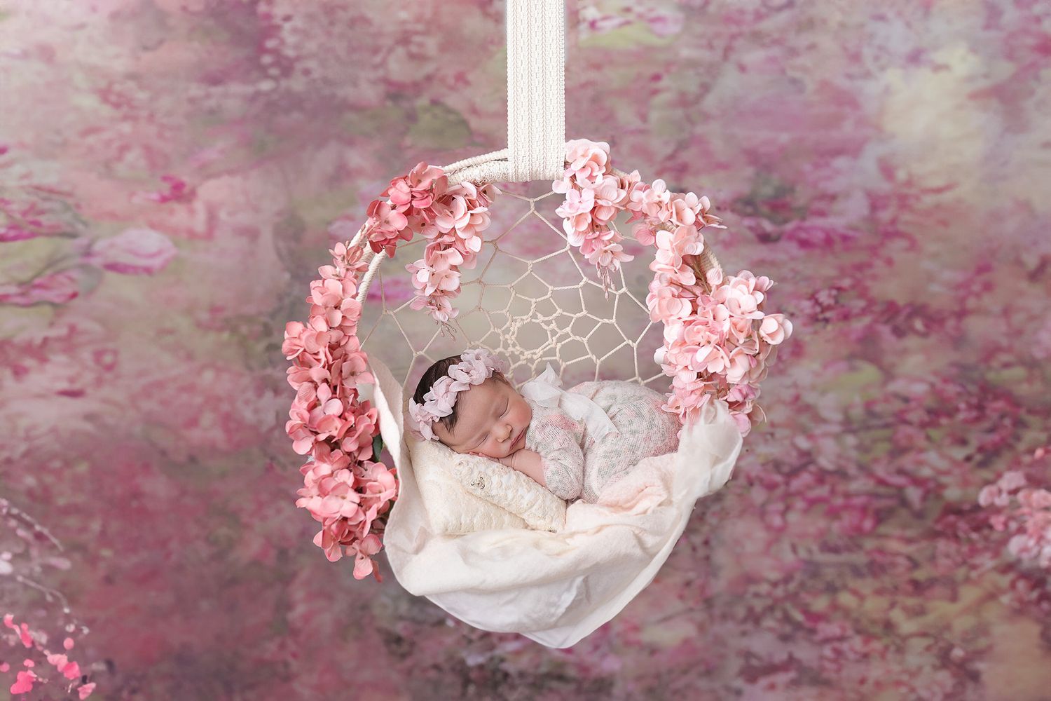 A white hanging basket decorated with pink cherry blossoms and draped fabric against a pink floral backdrop taken at Ana Brandt Baby Academy