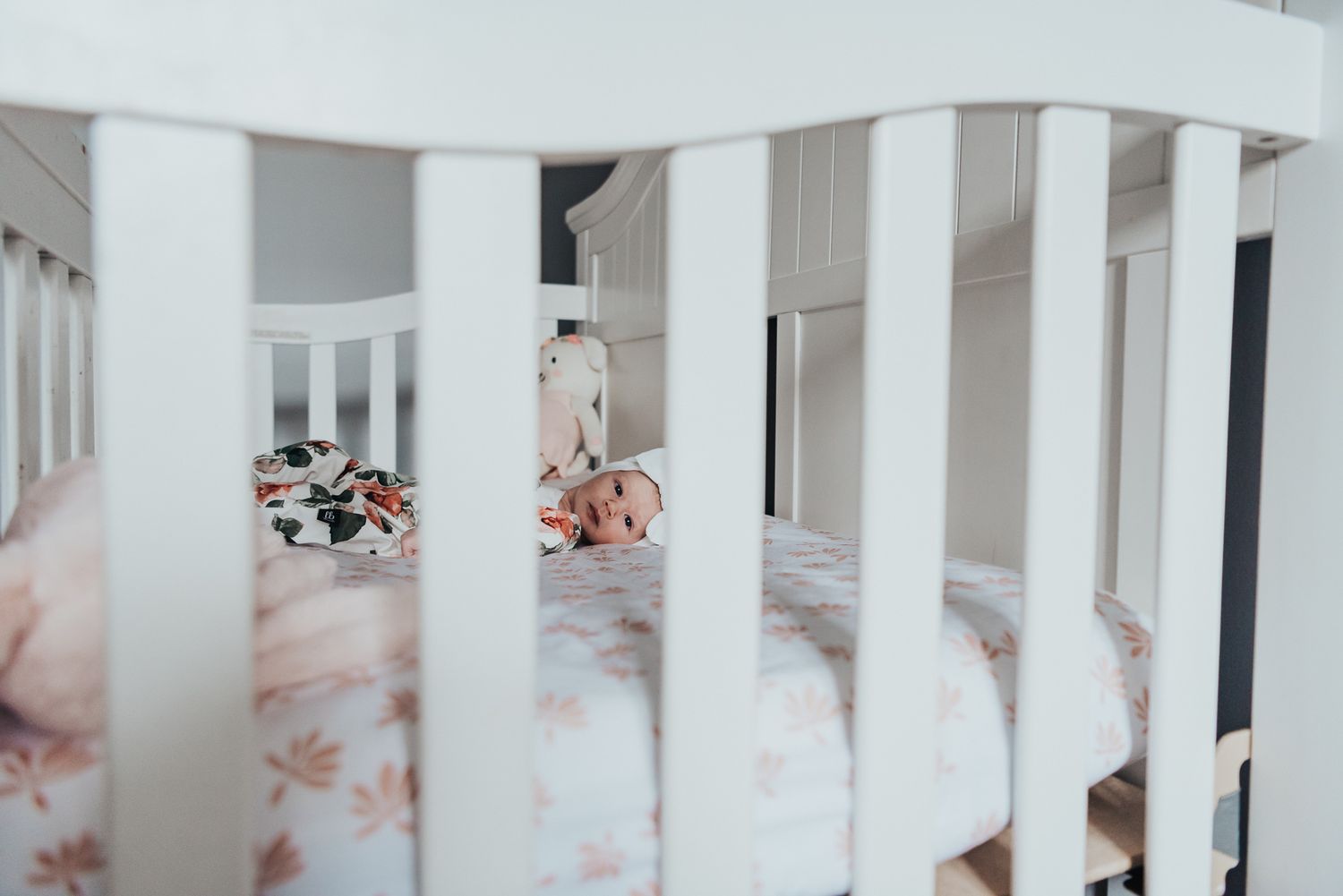 A little tiny newborn baby is seen through the posts on her crib as she lays looking toward the window.