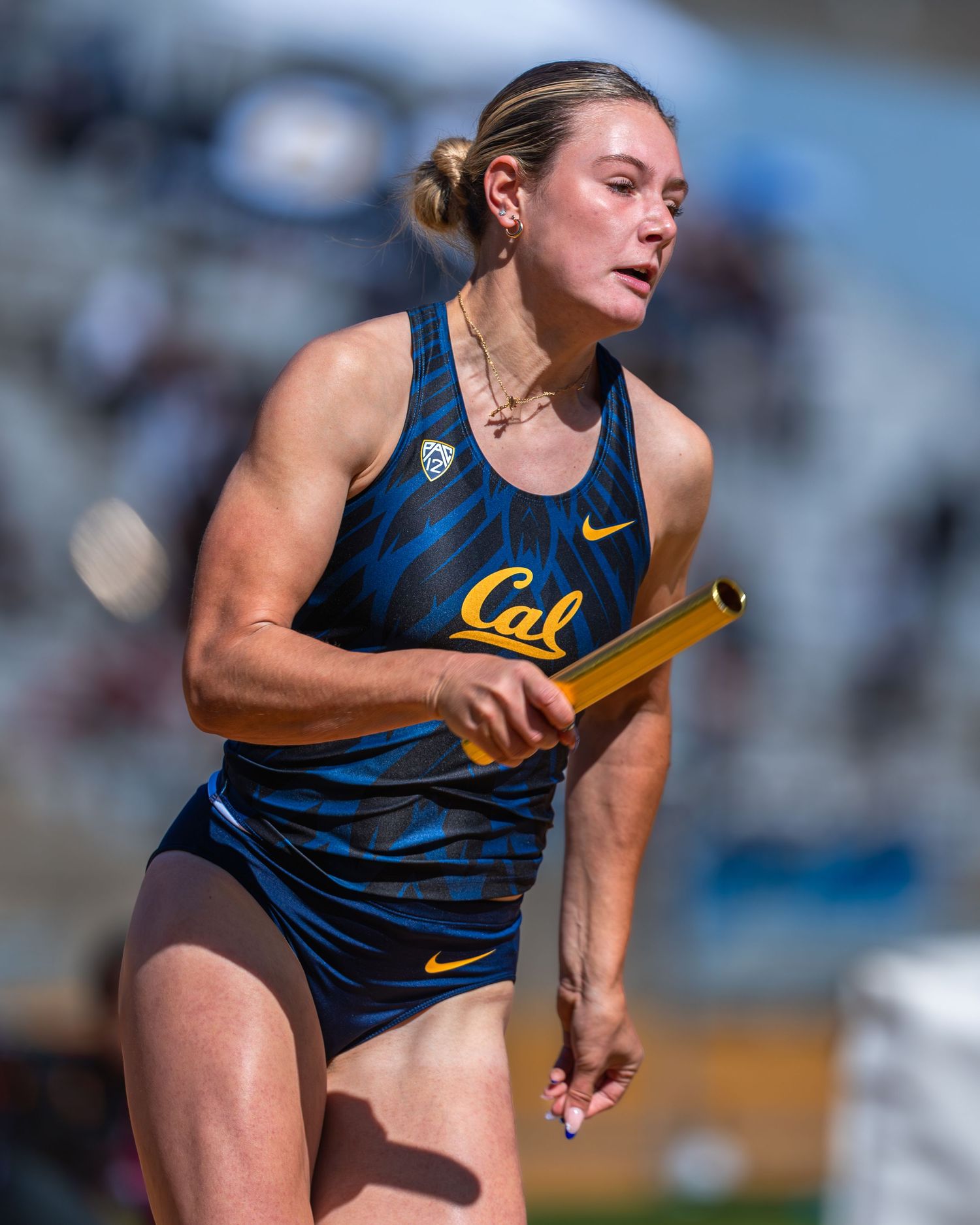 Track athlete in navy Cal uniform running with relay baton during competition.