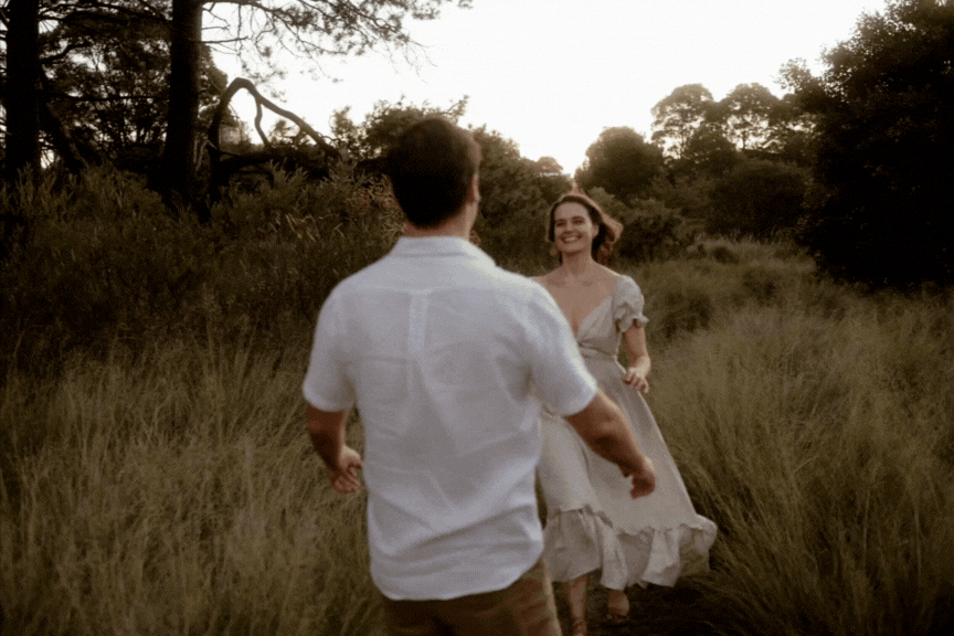 A couple in an open field shares a romantic moment at sunset as her white dress flows in the evening breeze.