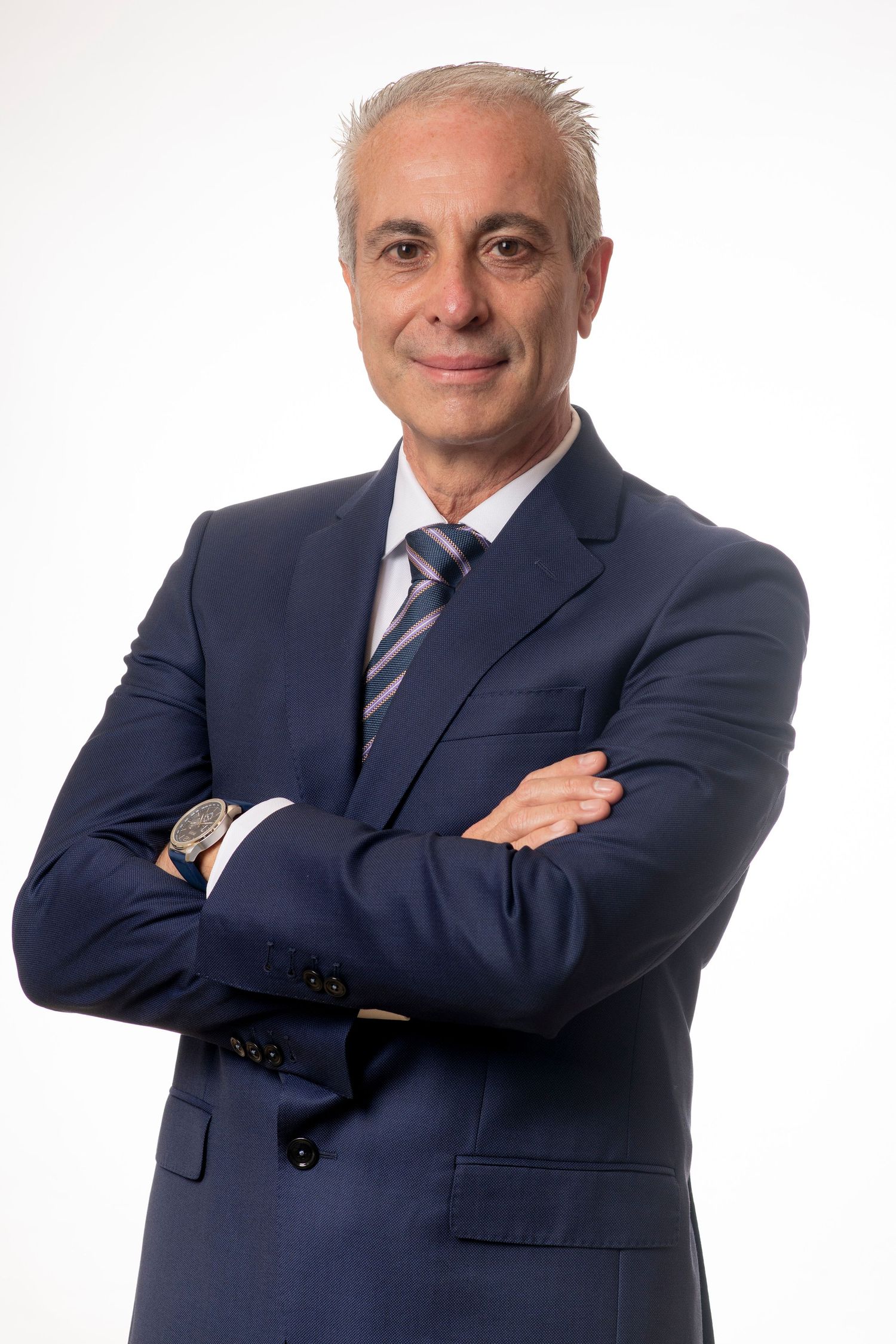 Foto profesional de alguien vestido con traje azul marino y corbata a rayas posando con los brazos cruzados sobre un fondo blanco.
