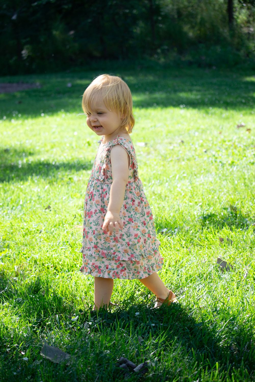 Small toddler in floral dress walking through sunny green grass.