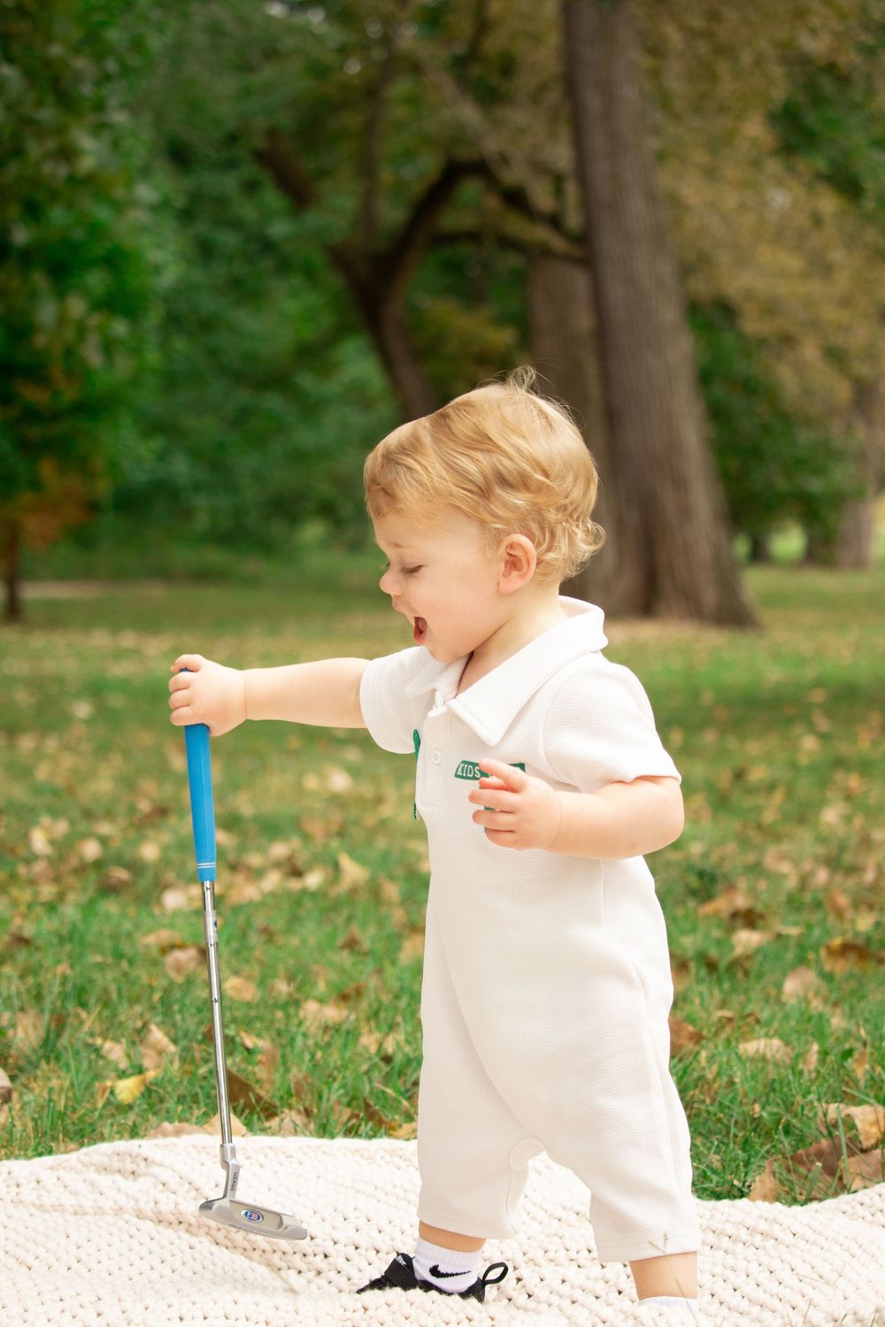 A toddler plays joyfully on a blanket surrounded by fallen leaves in an autumn park.