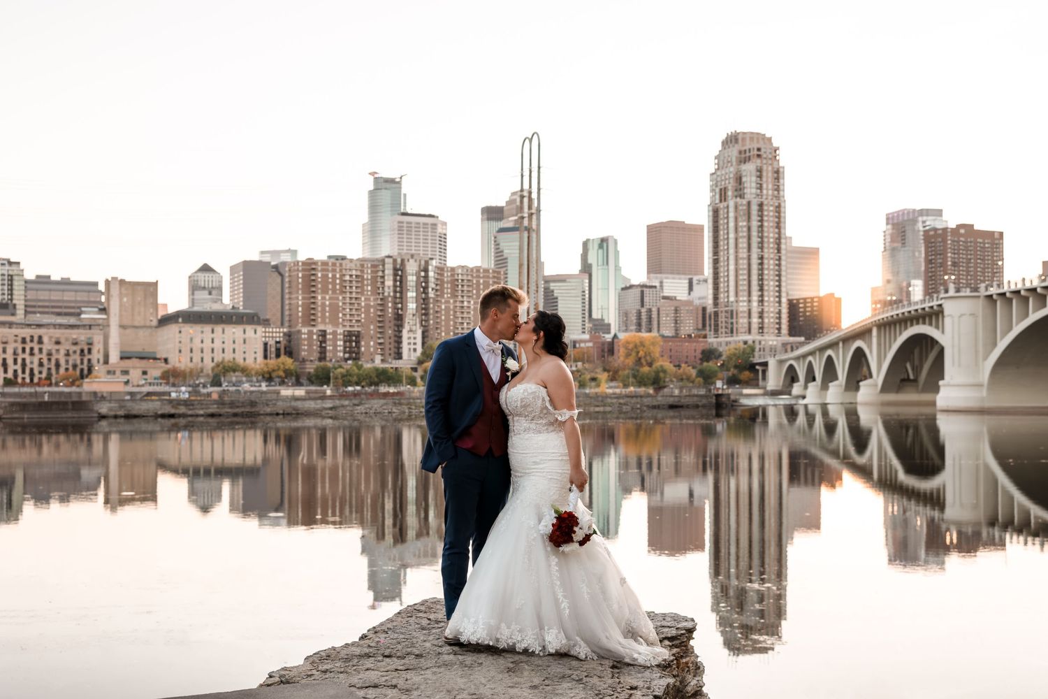 Wedding couple stands on riverfront with Minneapolis skyline and bridge reflection.