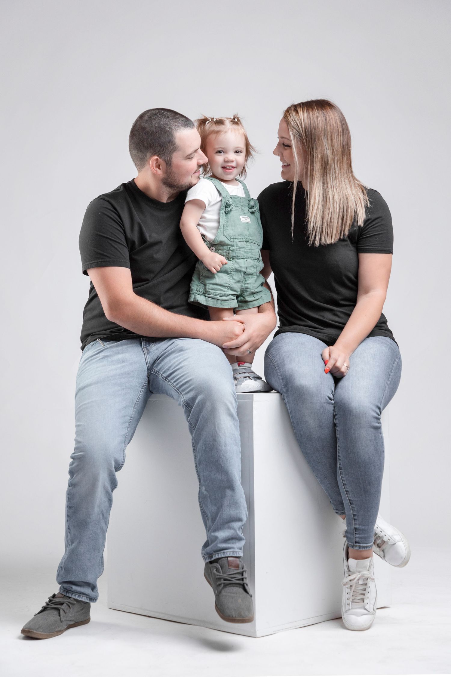 Family portrait with parents and toddler sitting together on white cube.