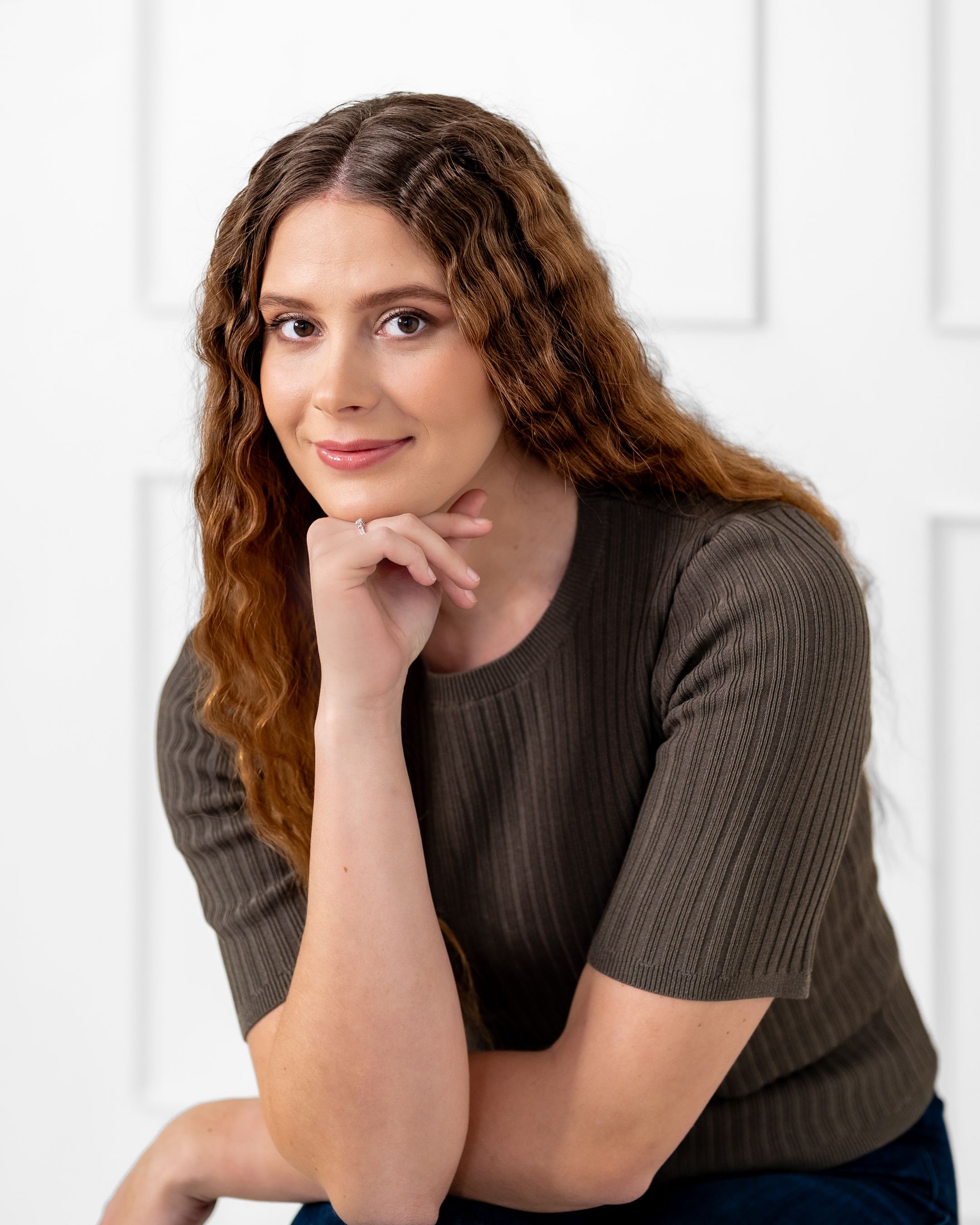 Casual portrait with long curly hair and brown ribbed top against white paneled wall.