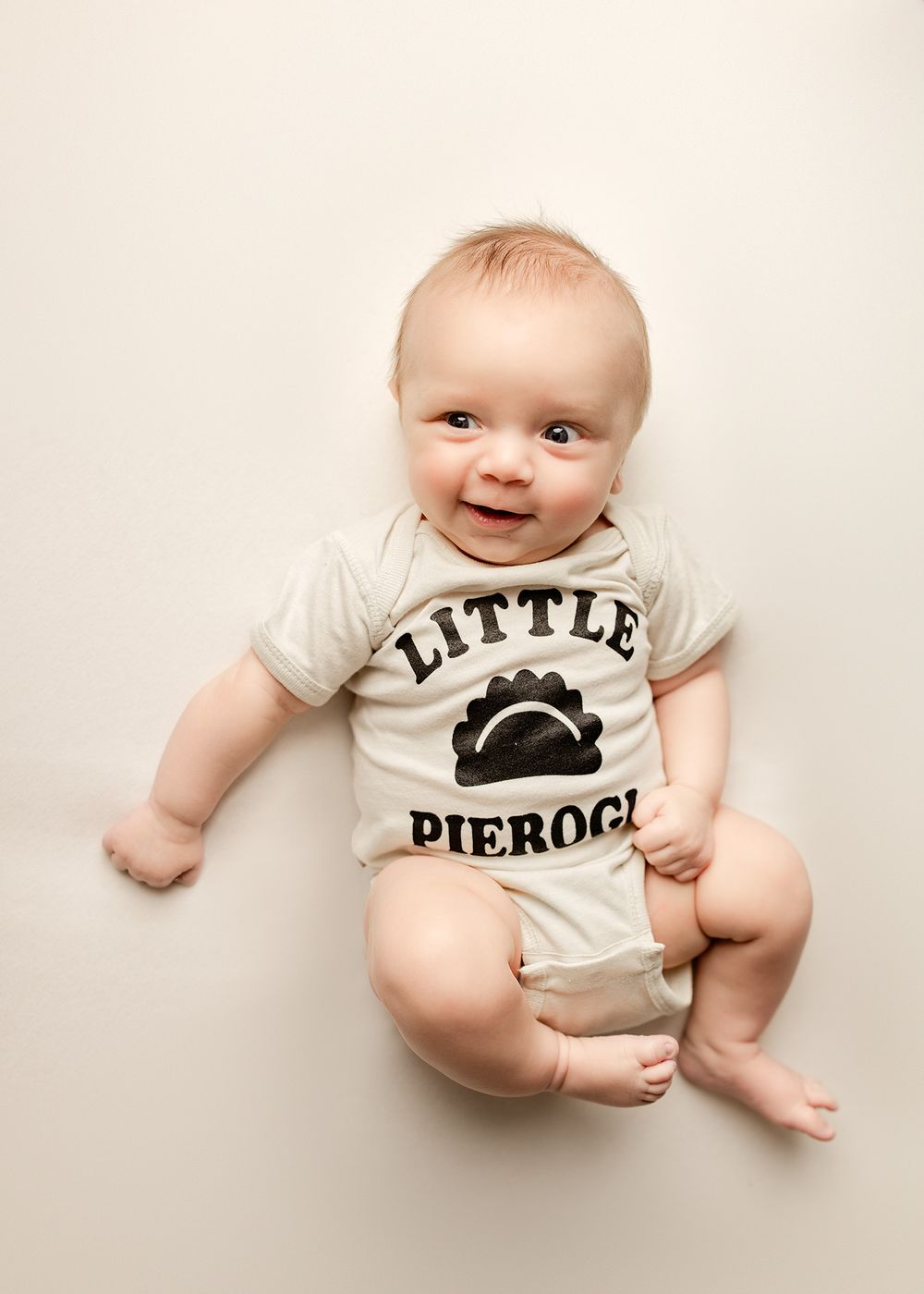 A smiling baby wearing a white Little Pierogi onesie lies on a cream background in a bright studio photo.