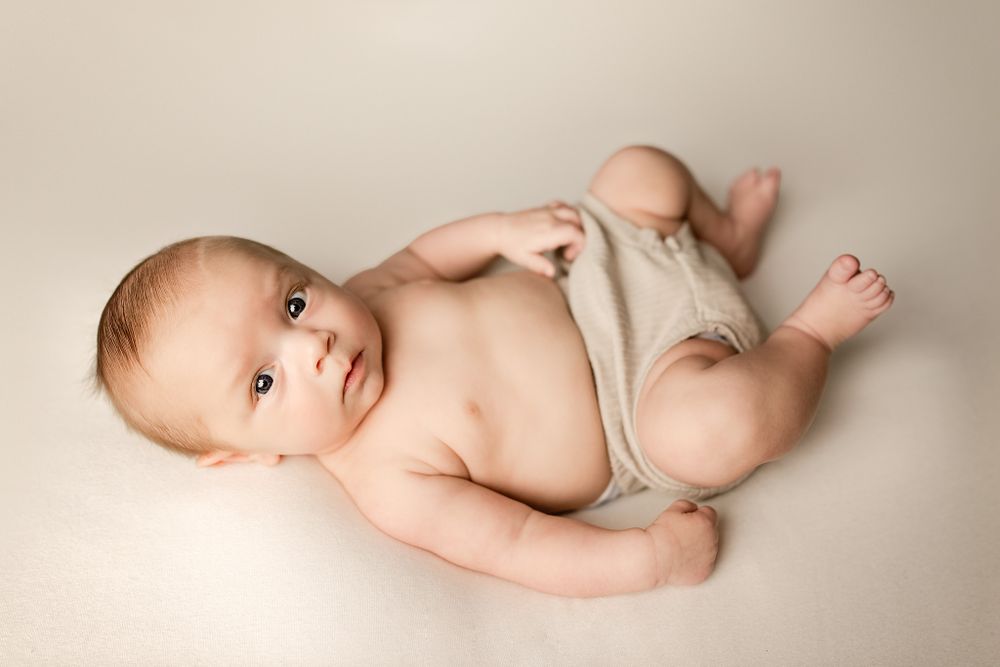 An adorable baby in a beige romper lies on their back on a cream blanket, looking up at the camera.