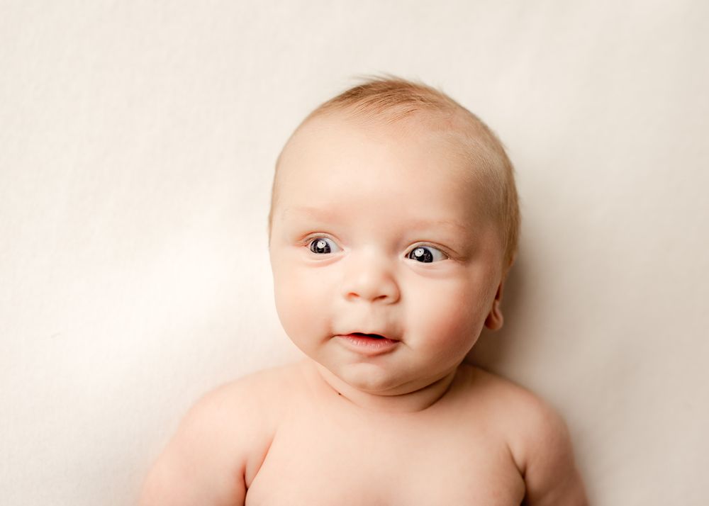 A close-up portrait of a wide-eyed baby lying on a cream blanket, looking up at the camera with a slight smile.