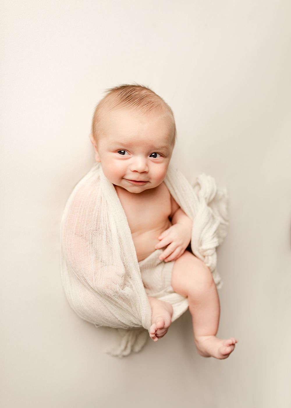 A smiling baby wrapped in a white muslin cloth sits upright on a cream background in a studio photo.