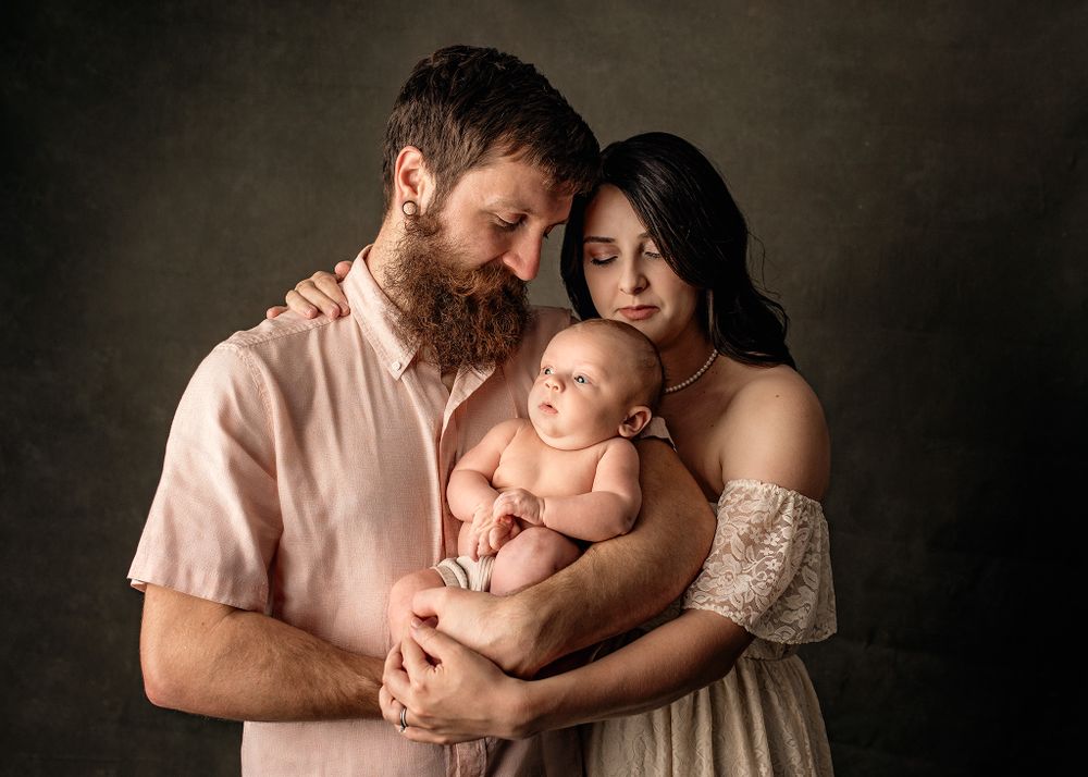 A bearded father and mother in a lace dress tenderly hold their newborn baby together in a studio portrait.