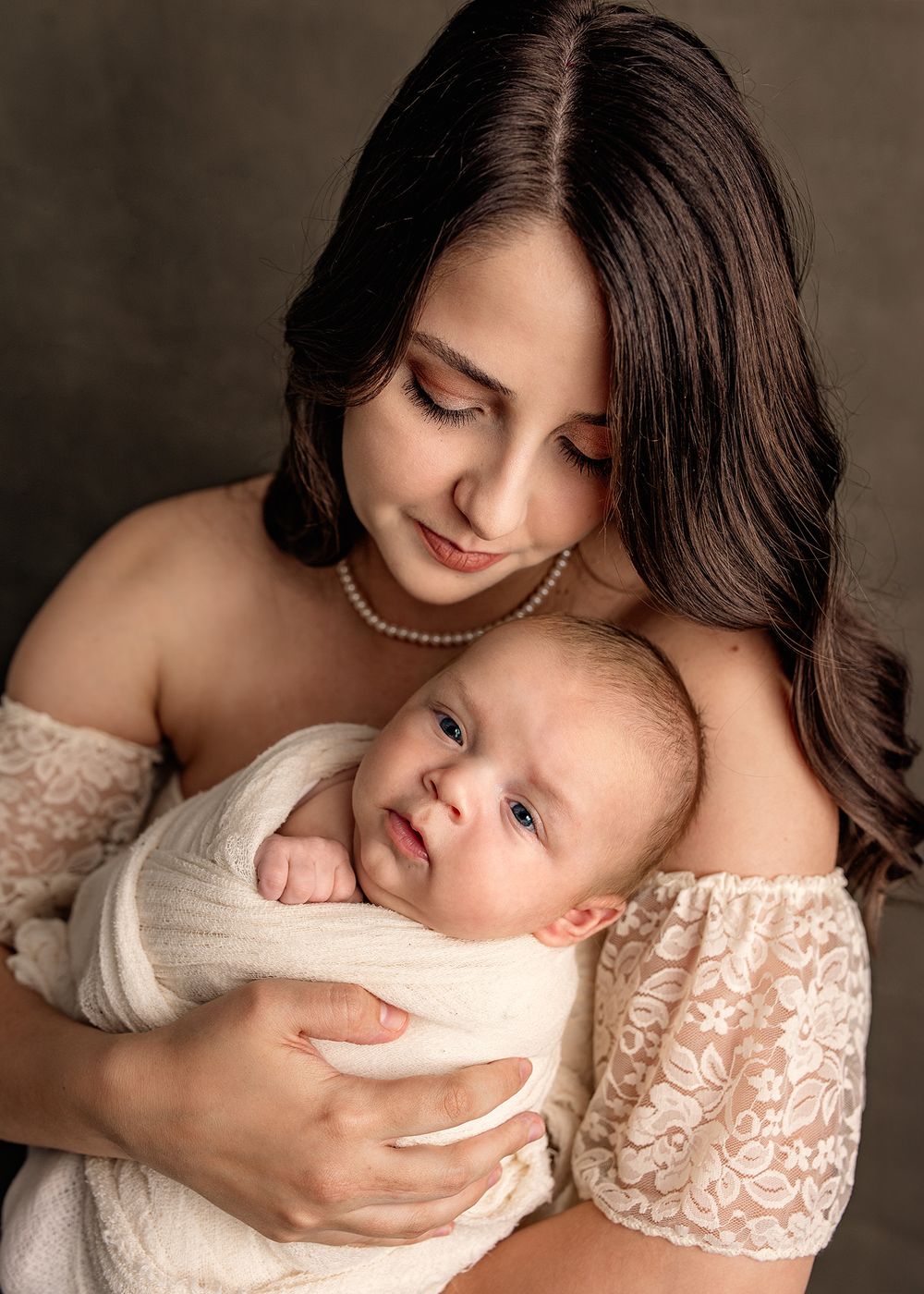 A brunette mother in a white lace dress holds her swaddled newborn baby close to her chest in a studio portrait.