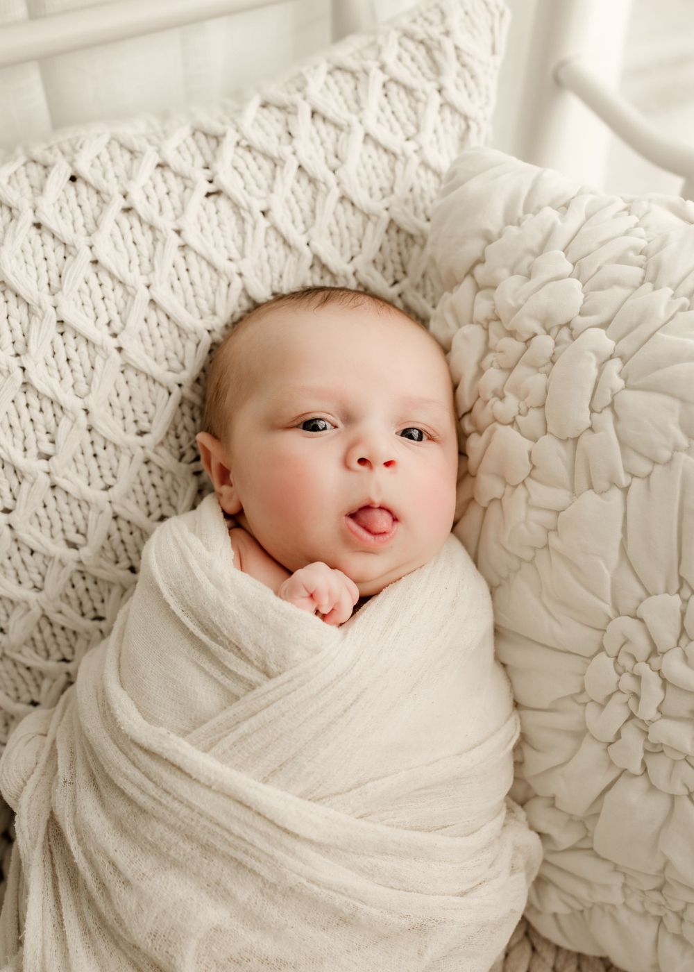 A swaddled newborn baby sticks out their tongue while lying in a white iron crib with knit blankets.