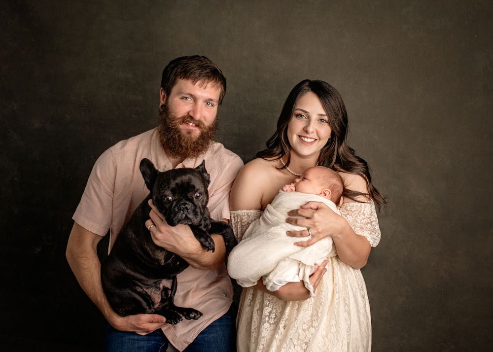 A bearded man and brunette woman pose with a black French Bulldog and newborn baby in a professional studio portrait.