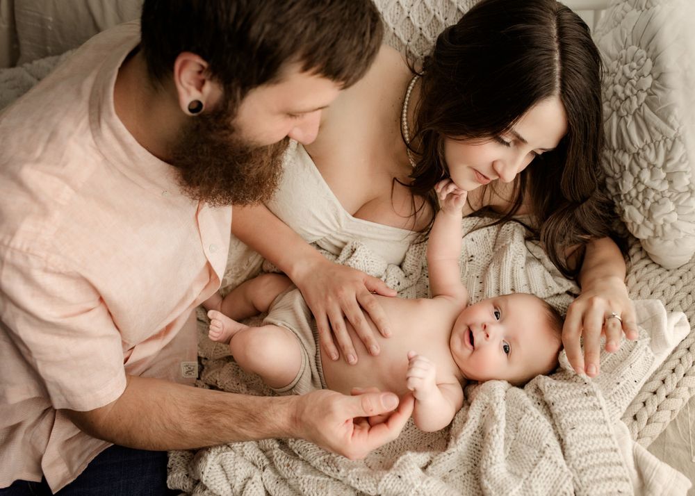 A bearded father and dark-haired mother smile and interact with their laughing baby on a white knit blanket.