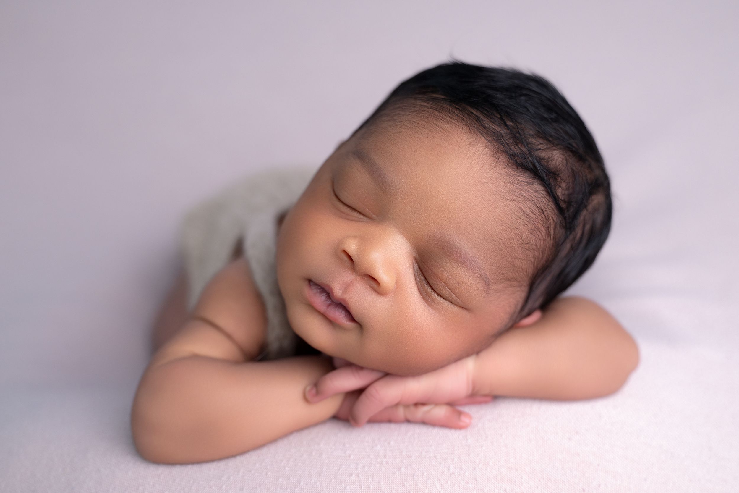 A peaceful newborn baby sleeping with hands tucked under chin on a soft lavender blanket.