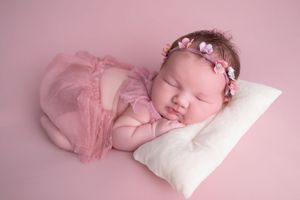 Newborn baby girl sleeping peacefully on white pillow, wearing pink tutu and floral headband, on pink background.