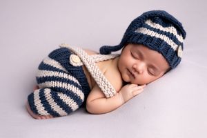 Sleeping newborn baby in navy striped knit hat and pants outfit, curled up peacefully on soft background.