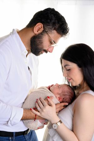 New parents lovingly gaze at their swaddled newborn baby, mother in white dress, father in white shirt. affordable photographer
