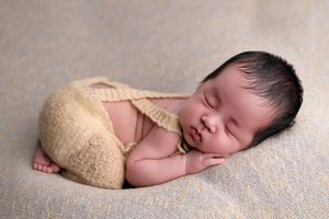 Peaceful newborn baby sleeping curled up wearing a knitted tan romper on a soft gray blanket.
