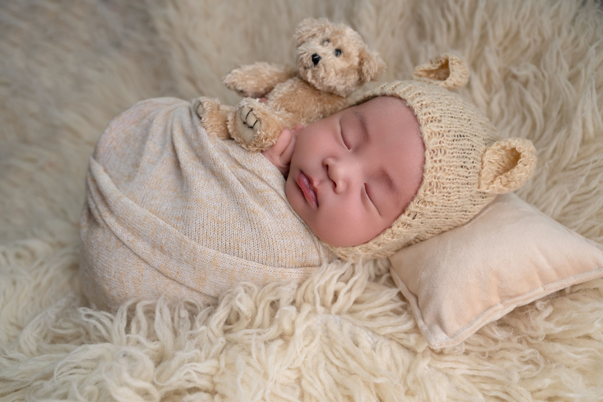 Sleeping newborn baby wrapped in cream knit swaddle wearing bear bonnet with teddy bear on fluffy white rug.