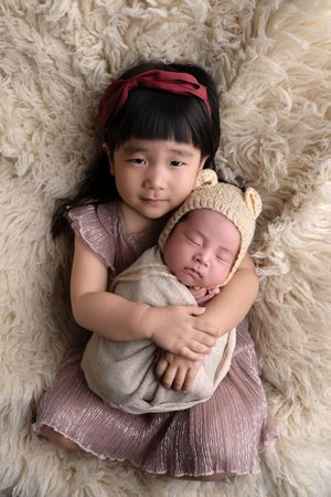 Young girl with red bow holds sleeping newborn sibling wrapped in cream swaddle with bear bonnet on fluffy rug.