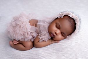 Sleeping newborn baby girl in pink ruffle outfit and bonnet on white knit blanket.
