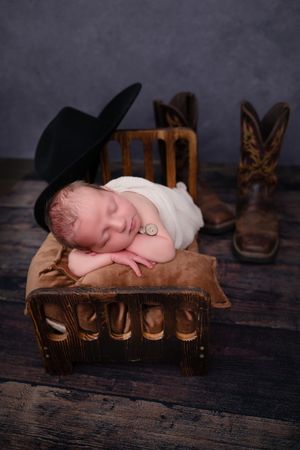 Newborn baby sleeping on tiny wooden bed with black cowboy hat and brown boots in rustic western-themed photo.