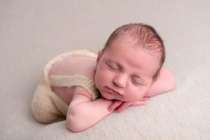 Newborn baby sleeping peacefully in a froggy pose wearing a cream knit outfit on a soft beige blanket.