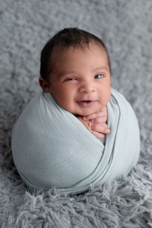 Smiling newborn baby wrapped in a soft blue swaddle blanket, posed upright on a gray fluffy rug.