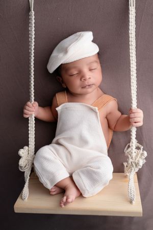 Sleeping newborn baby in white sailor outfit and cap resting on a wooden macrame swing.