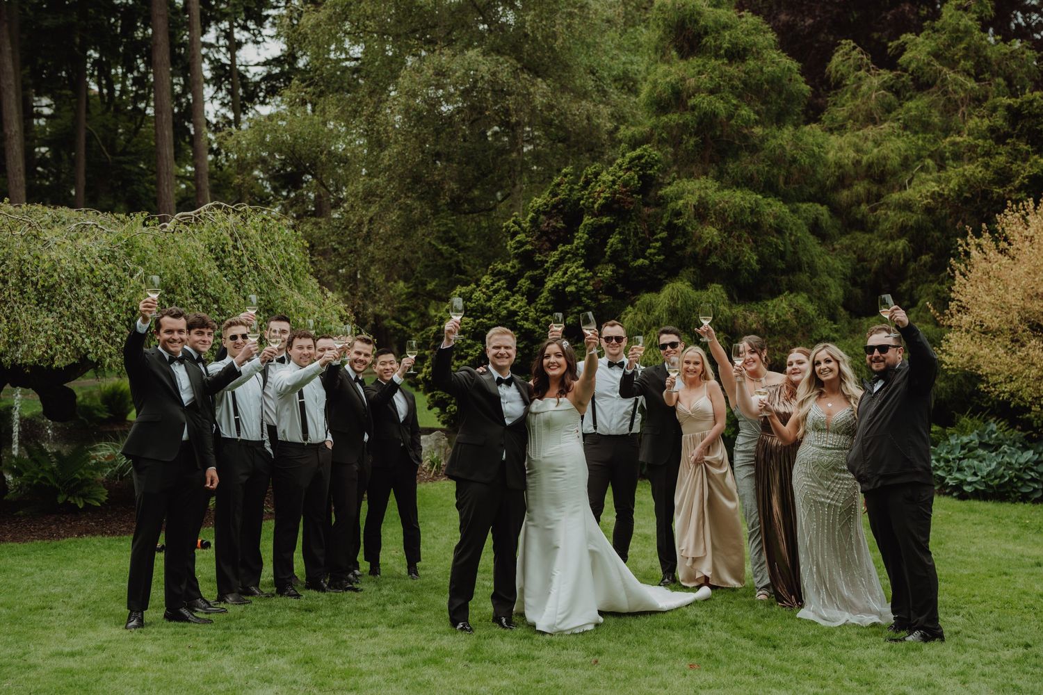 Joyful wedding party celebrates on a lush green lawn surrounded by tall trees, raising hands in celebration.