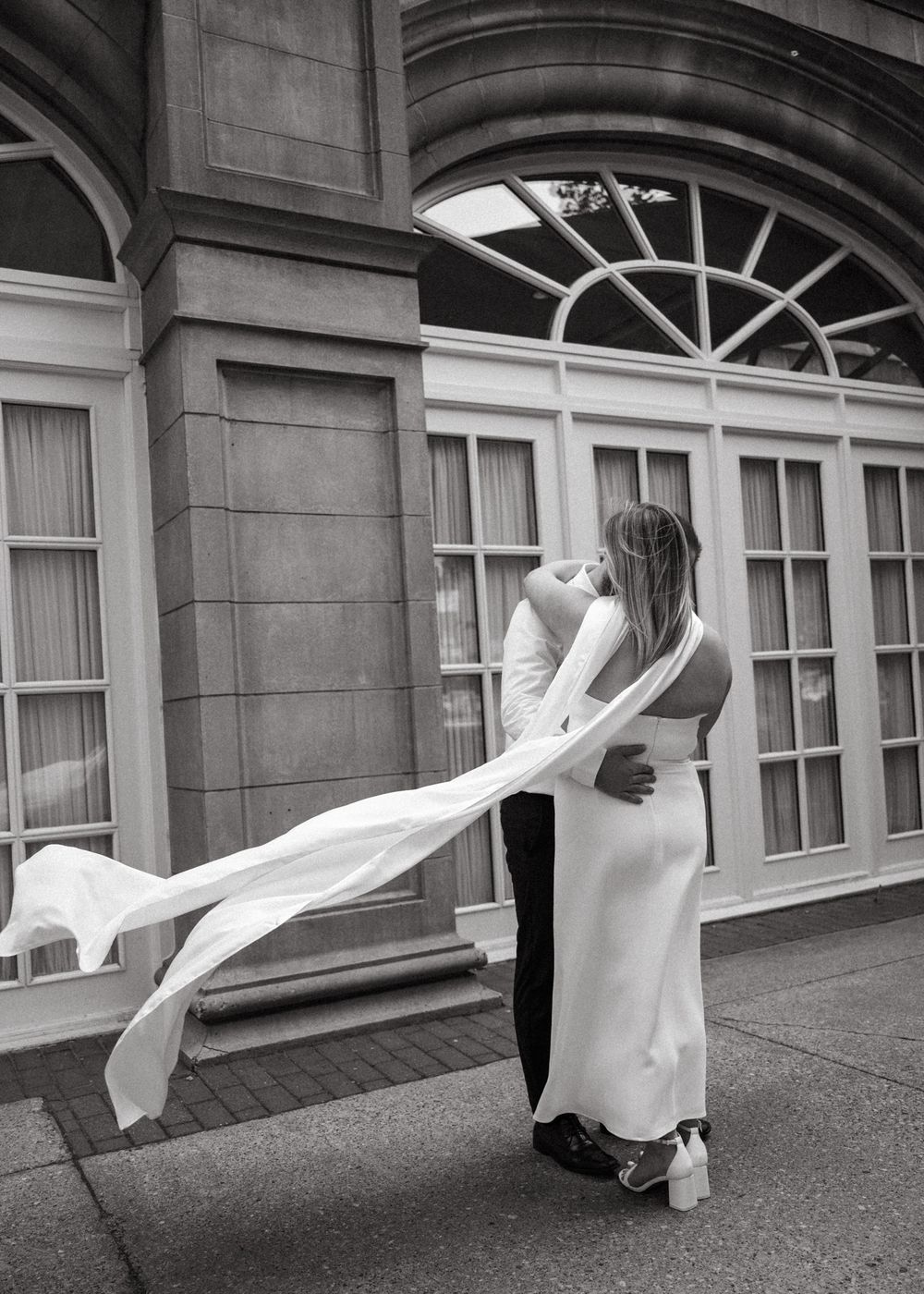 A romantic black and white wedding photo with a flowing veil caught in the breeze outside an elegant building entrance.