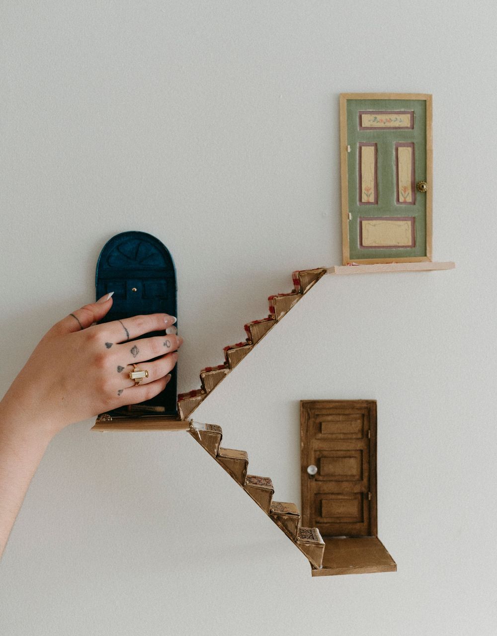 A hand opens a miniature wooden staircase door mounted on a white wall, showing a sequence of progressive movements.
