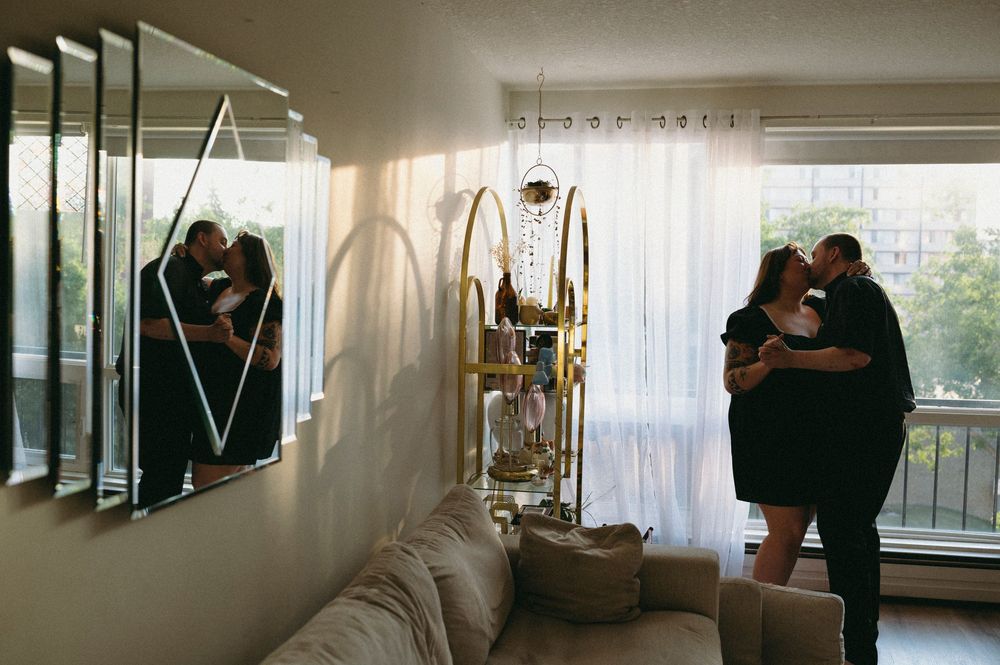 Two silhouetted figures embrace in a bright apartment interior with curtains and a ladder visible.