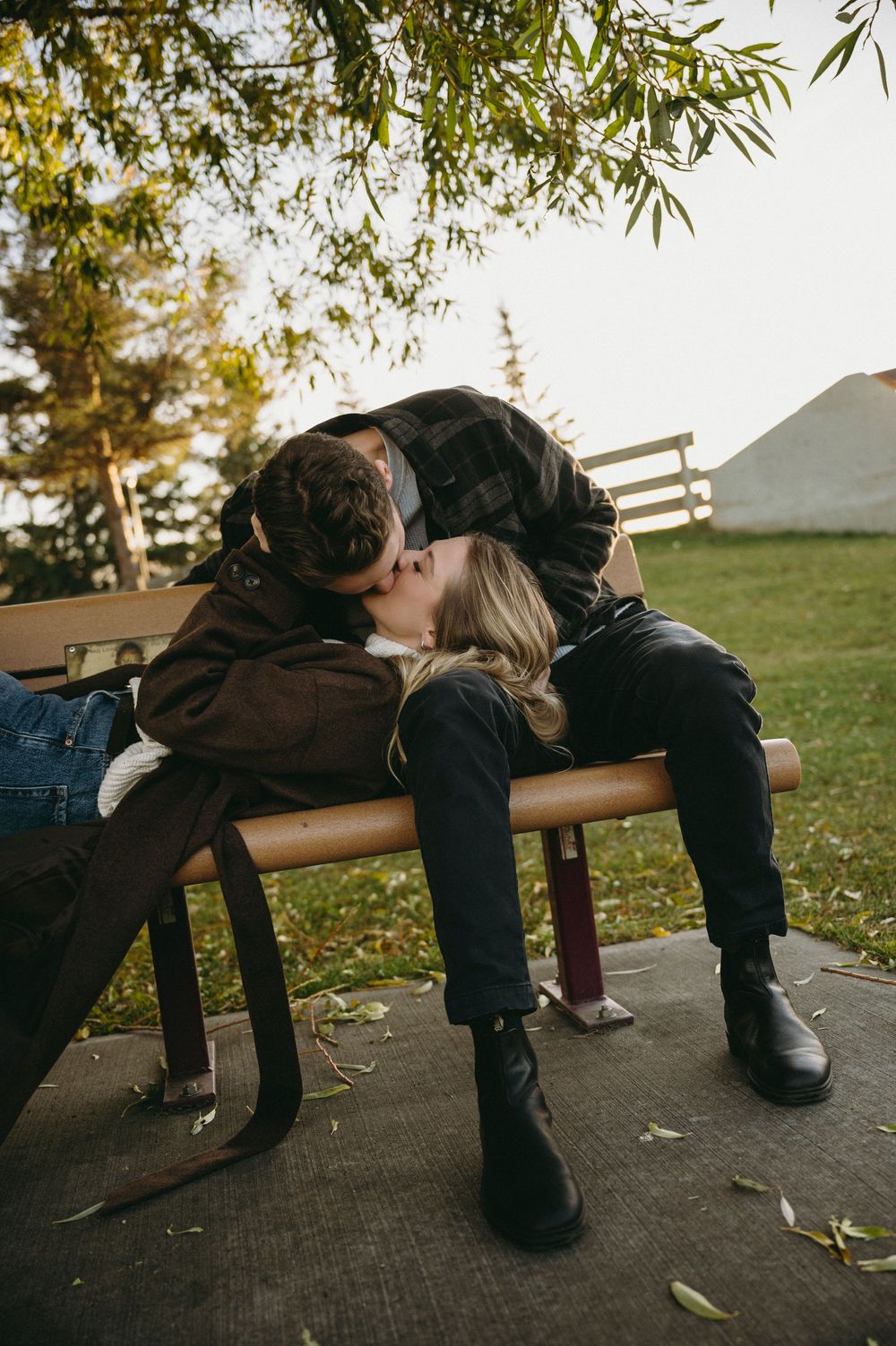 Two people rest together on a wooden park bench on an autumn day as leaves fall around them.
