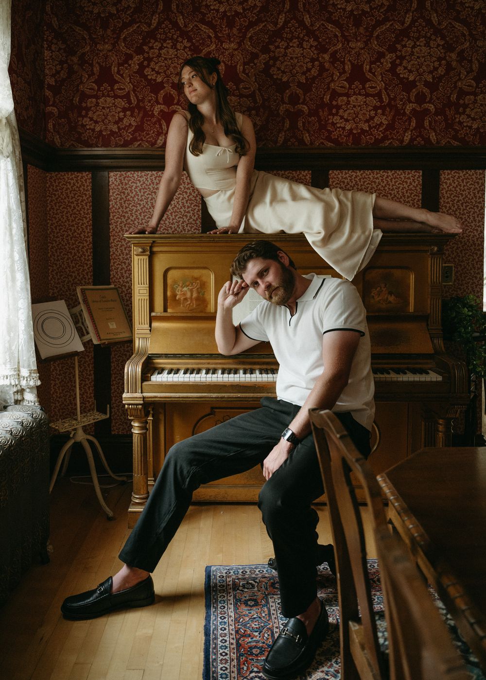 A person in white shirt and dark pants sits on a hardwood floor near a piano in a vintage-style room.