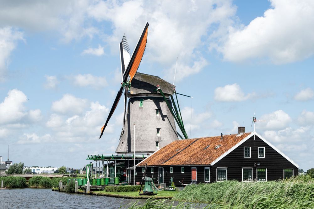 Classic Dutch windmill with large sails stands beside a traditional farmhouse on the waterfront.