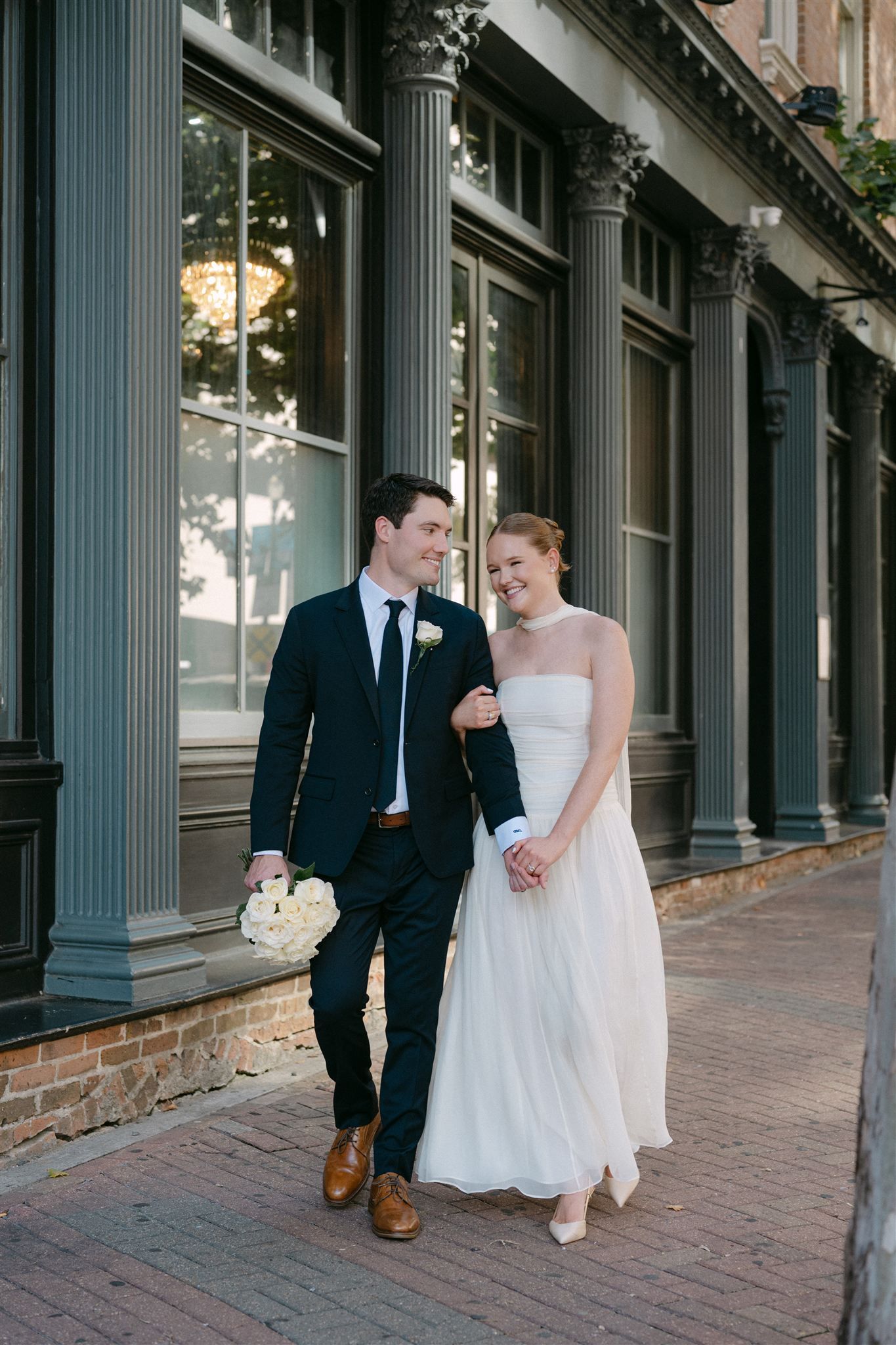 A couple in formal attire smiles and interacts on a brick sidewalk outside a building with classical columns.