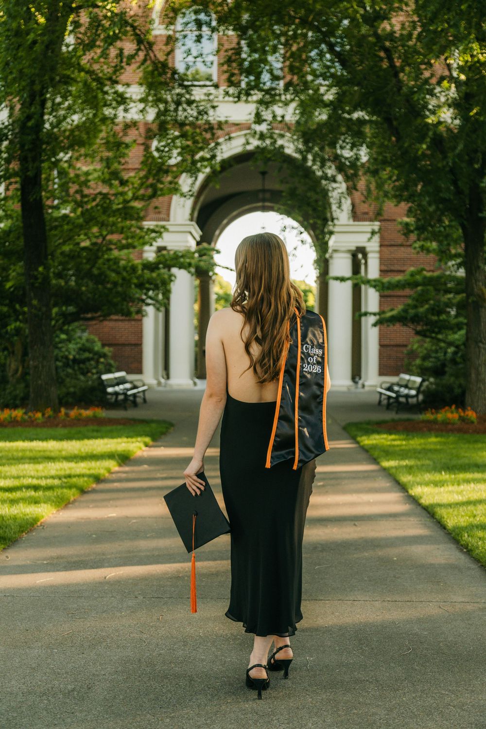 Woman in black dress holding graduation cap walks toward brick archway on tree-lined campus path.